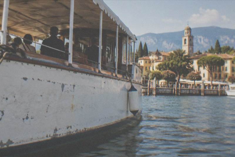 Weathered white passenger boat with canopy and seated people in the foreground, Lake Garda lakeside town, church tower, trees and mountains beyond.