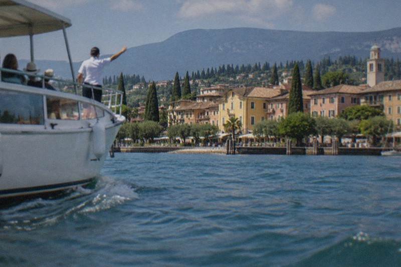 A white canopy boat moves on blue water as a man in a white shirt points toward a Lake Garda lakeside town of pastel buildings, cypresses and mountains.