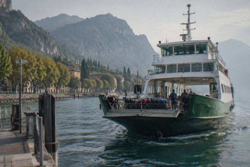 A green-and-white ferry with people and bikes on the open stern nears a wooden dock, mountains and trees along the shore behind, on Lake Garda.