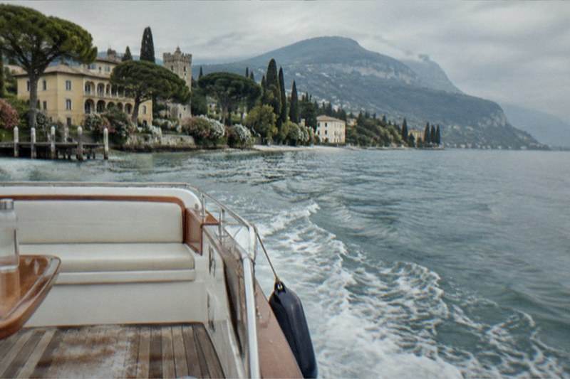 From a boat's wooden deck with white cushions and railing, a wake trails toward Lake Garda lakeside villas, pines and mountains under a cloudy sky.