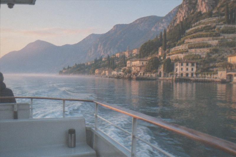 From a boat stern, a person stands by the rail as the wake trails toward pale Lake Garda lakeside houses and terraced slopes under mountains.