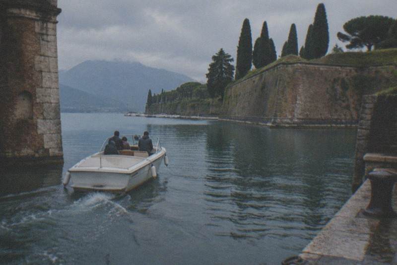 A small white motorboat with several people heads away through a canal between stone walls, cypress trees and a mountain under gray clouds, on Lake Garda.