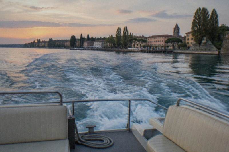 From a boat stern, empty padded seats, a coiled rope and railing face a foamy wake toward a tree-lined Lake Garda lakeside town at sunset.