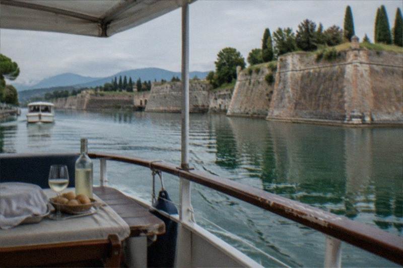 From a boat deck, a table with wine bottle, glass and bread faces a stone fortress along calm water under an overcast sky, on Lake Garda.