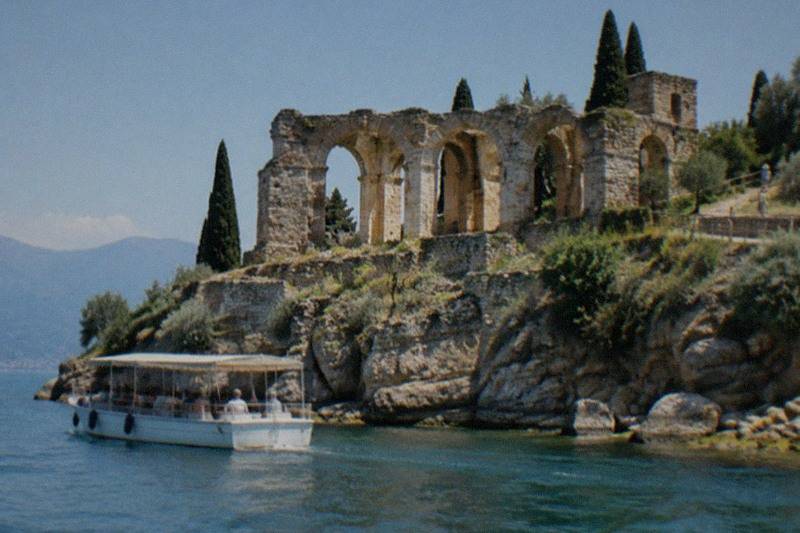 A white tour boat sits on calm water below arched stone ruins on a rocky headland with cypress trees, on Lake Garda.