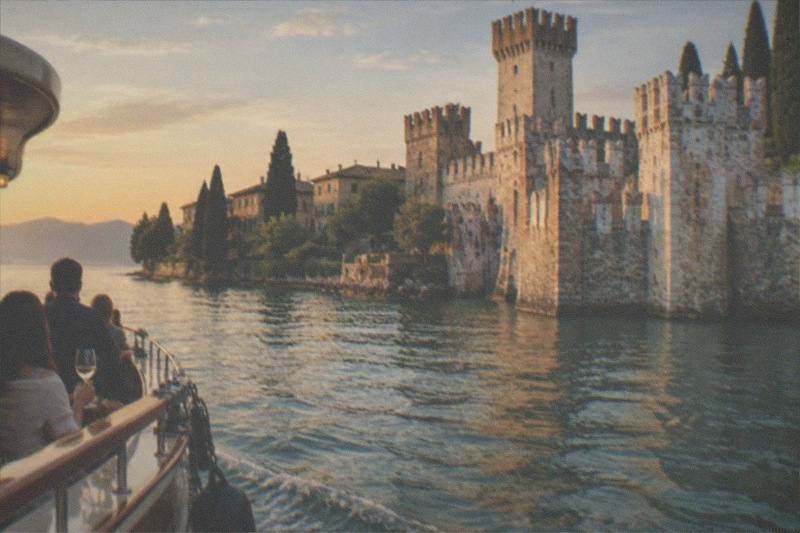 From a boat deck, people look toward a stone Lake Garda lakeside castle with towers at dusk; one passenger holds a wine glass.