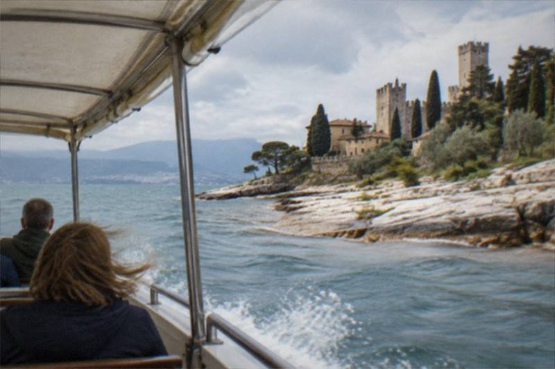A low tour boat drifts close beneath the rocky edge of the Sirmione peninsula, its passengers looking up at the overgrown cliffs and villas that stand above the waterline.