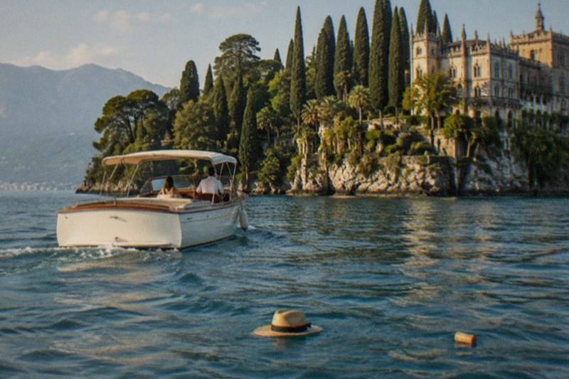 A white canopy motorboat with two people heads toward a villa on a cypress-covered rocky island; a straw hat floats in the foreground, on Lake Garda.