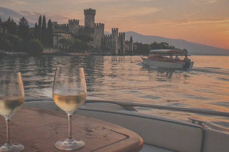 Two glasses of pale wine sit on a wooden table aboard a boat, with a motorboat passing a Lake Garda lakeside castle under an orange sunset sky.
