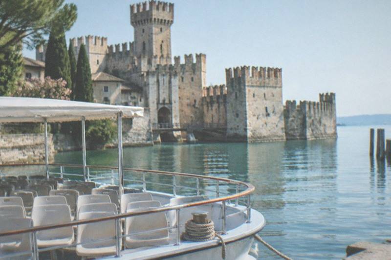 An empty white tour boat with a canopy sits on calm water before a stone castle with towers and battlements under a clear sky, on Lake Garda.