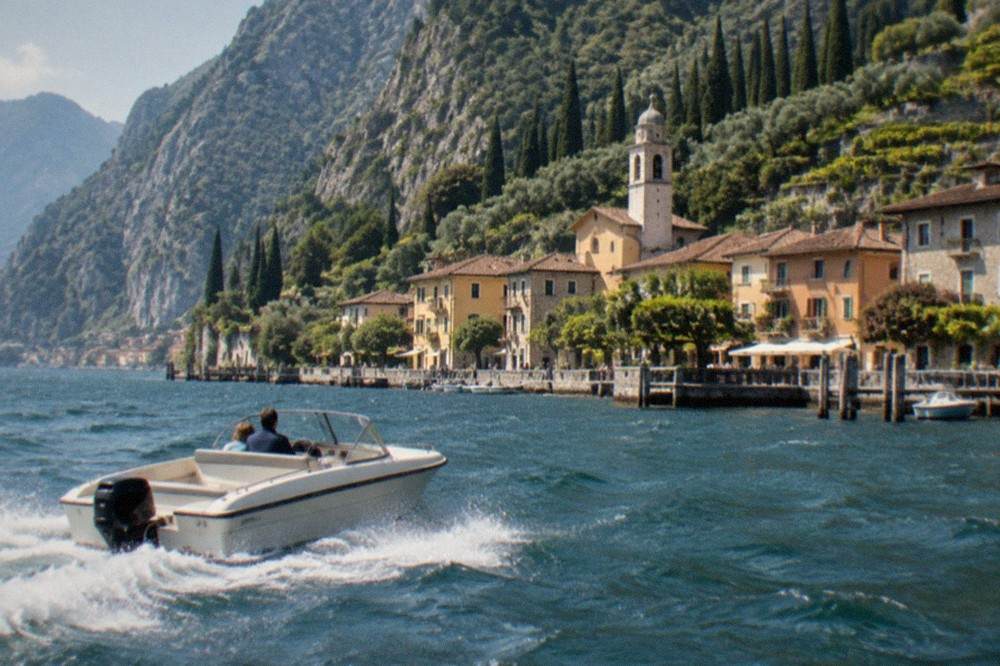 White motorboat with two people on Lake Garda, heading toward a village of ochre buildings and a church tower beneath steep wooded mountains.