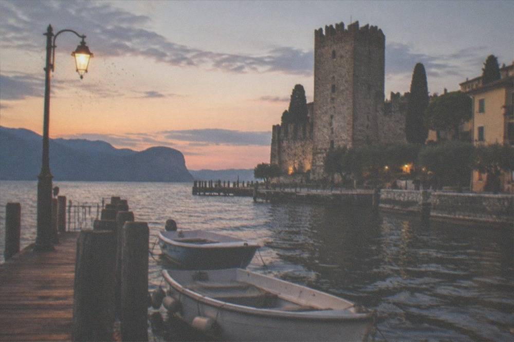 A wooden jetty with small boats moored along it and a castle tower across the water at dusk
