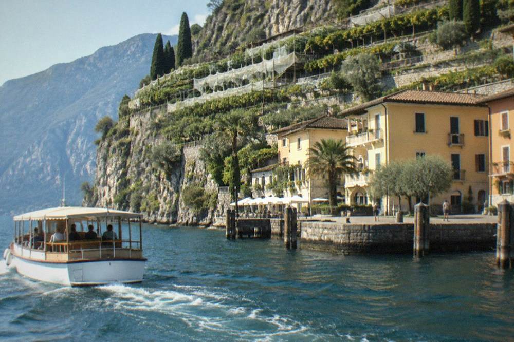 A cruising boat with a shaded deck passing olive groves and a small lakeside village on Lake Garda