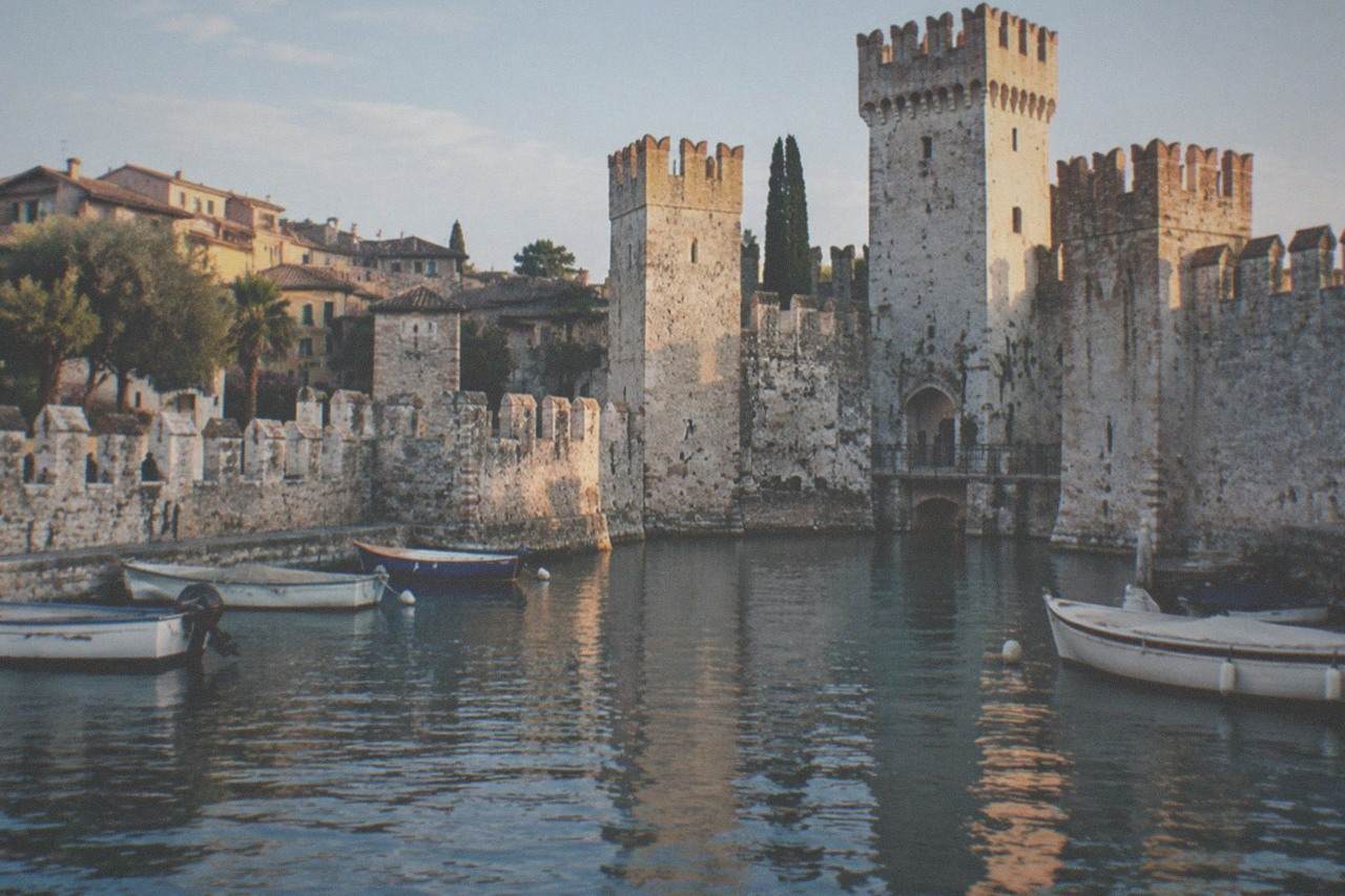 A stone castle with crenellated walls rising from the edge of a lake, with small boats moored in the foreground and a town of tiled roofs behind.