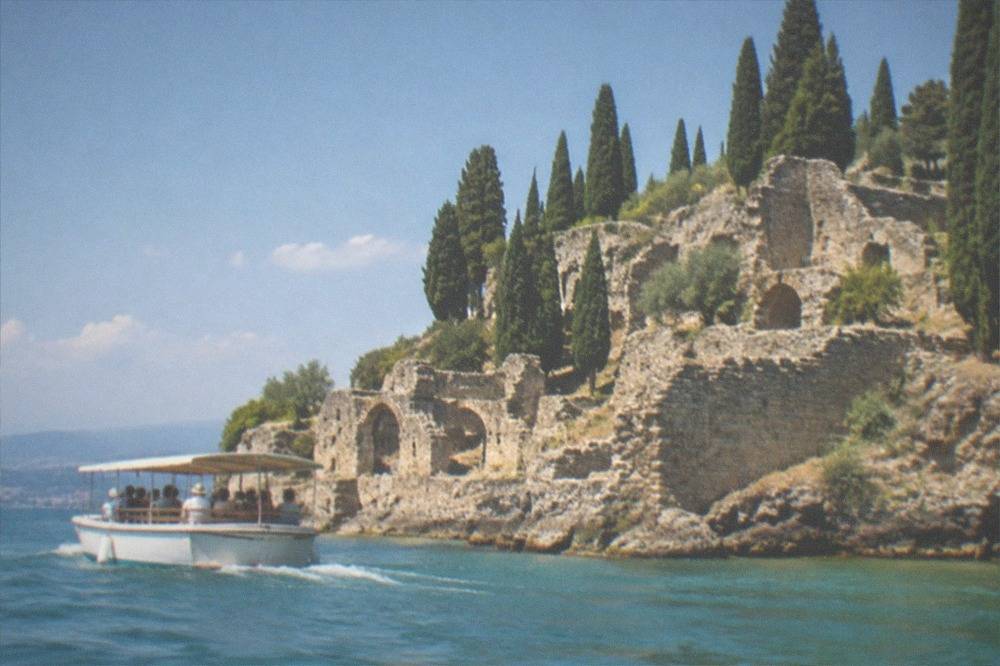 A low boat passing close to a rocky peninsula with cypress trees and old stone walls