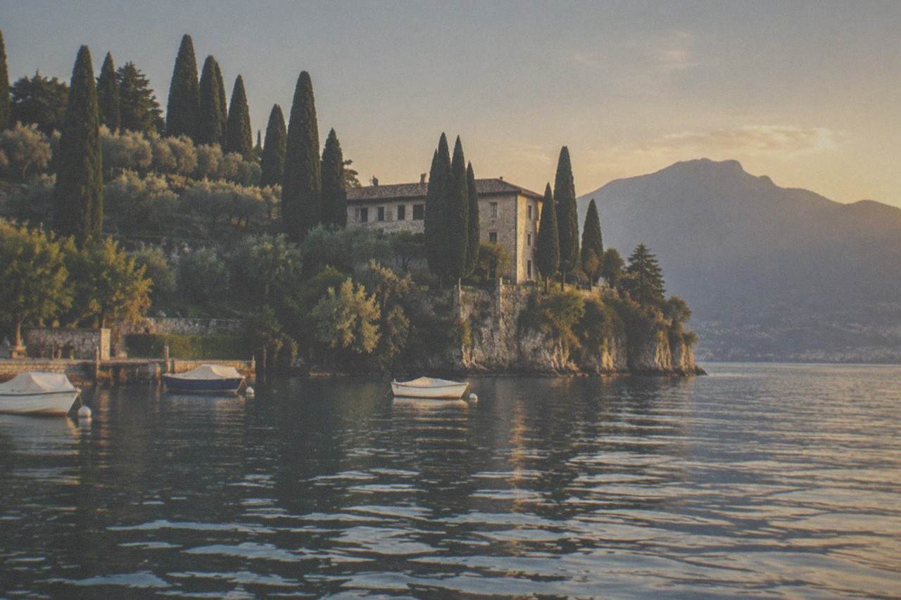 Stone villa on a cypress-lined cliff above a calm lake, with three small boats moored and mountains in the distance, on Lake Garda.