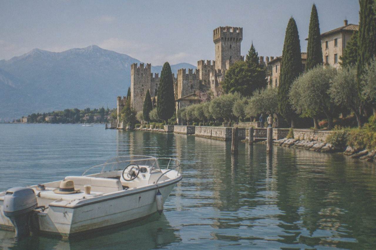 A white motorboat sits on calm water before a stone castle with battlements and cypress trees, mountains across Lake Garda.