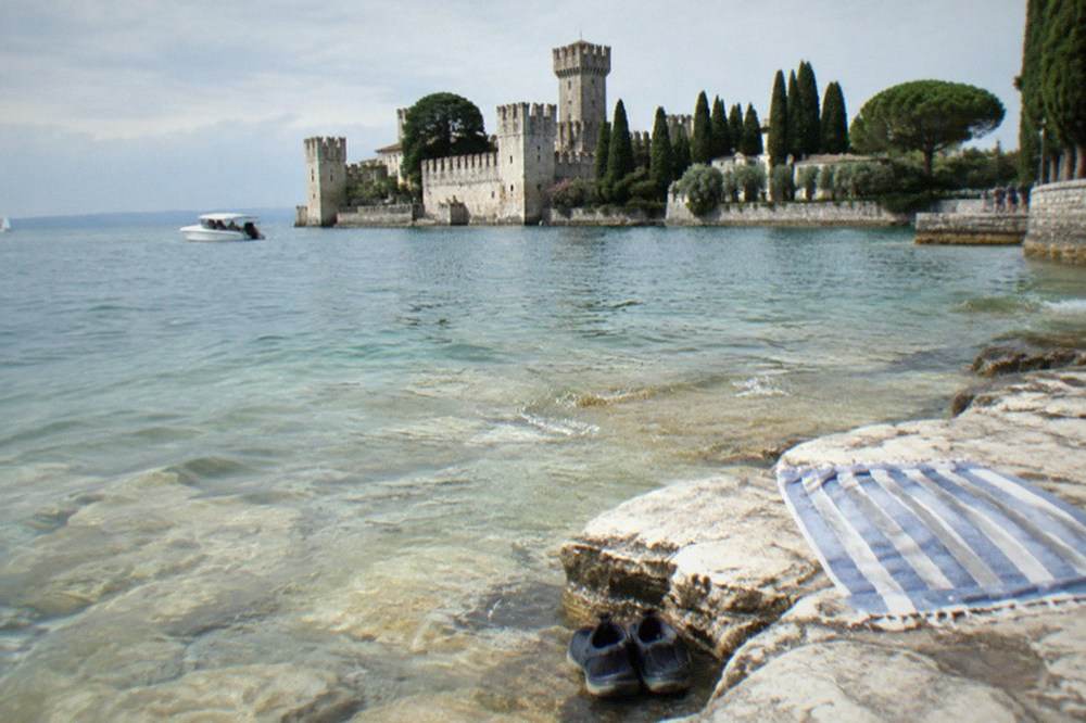 Dark shoes and a striped towel on pale rocks at the water's edge, with a Lake Garda lakeside stone castle, trees and a small white boat beyond.