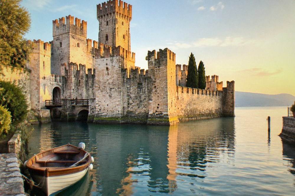A wooden boat moored at a stone pier with a castle tower rising above a lakeside old town