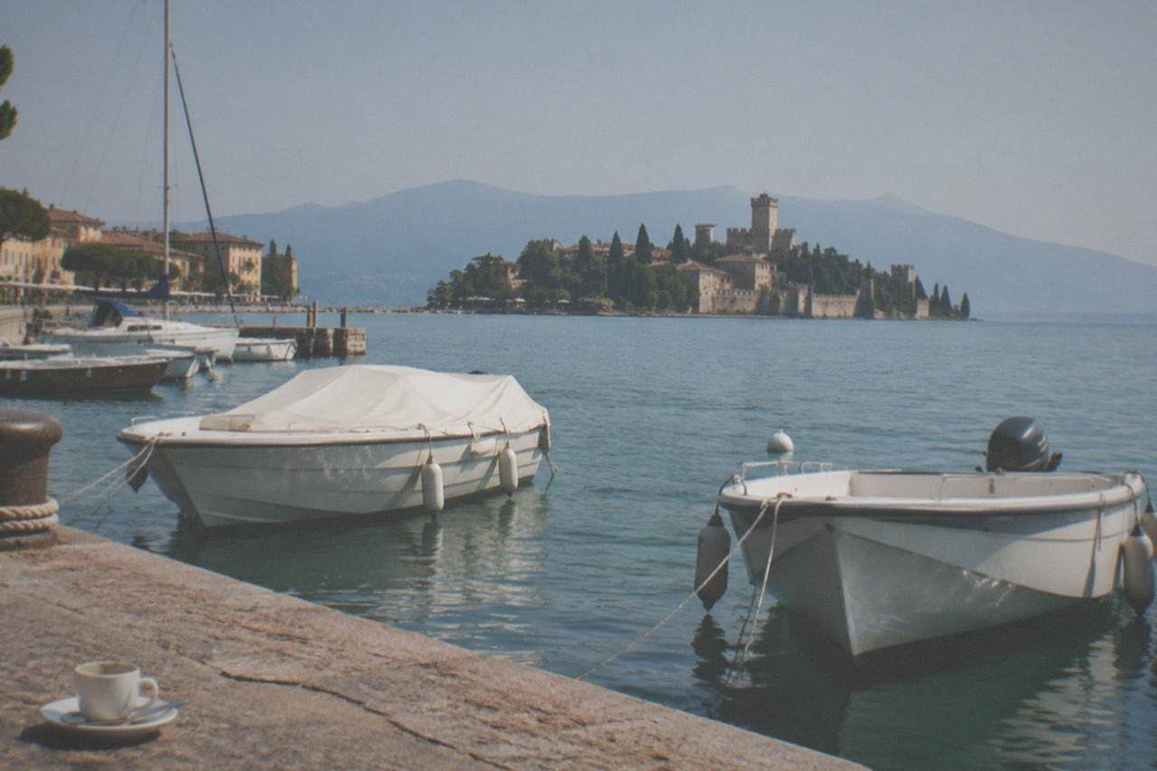 Two Lake Garda waterfront towns seen across calm water with moored boats and distant hills