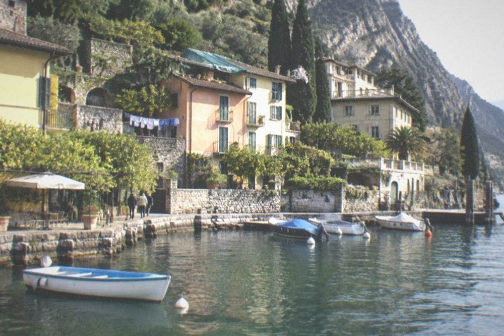 Lakeside village with pastel houses, a stone quay, and small boats moored in calm water beneath a steep wooded hillside, on Lake Garda.