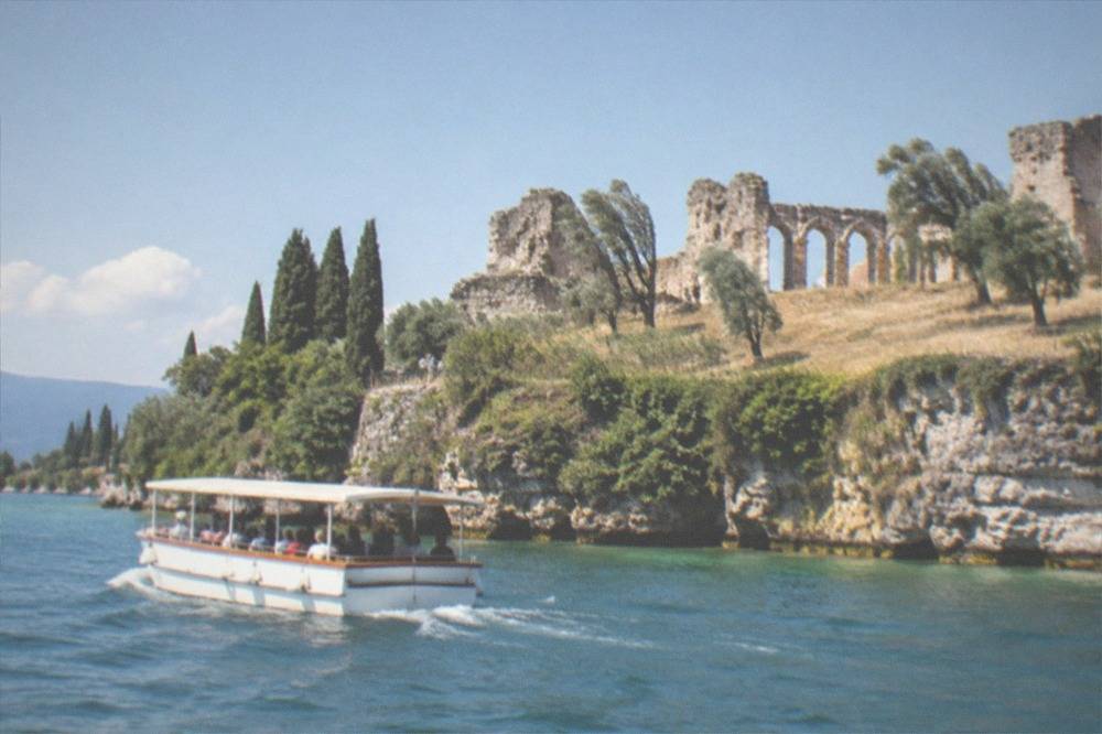 A tour boat passing the low cliffs and olive groves of the Sirmione peninsula on a bright afternoon