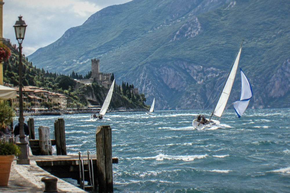 Sailboats heel on choppy Lake Garda water beside a wooden pier, with a hillside castle, lakeside houses and mountains beyond.