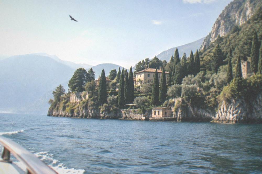 From a boat, a rocky forested lakeshore with cypresses and a hillside villa, a bird overhead and mountains beyond, on Lake Garda.