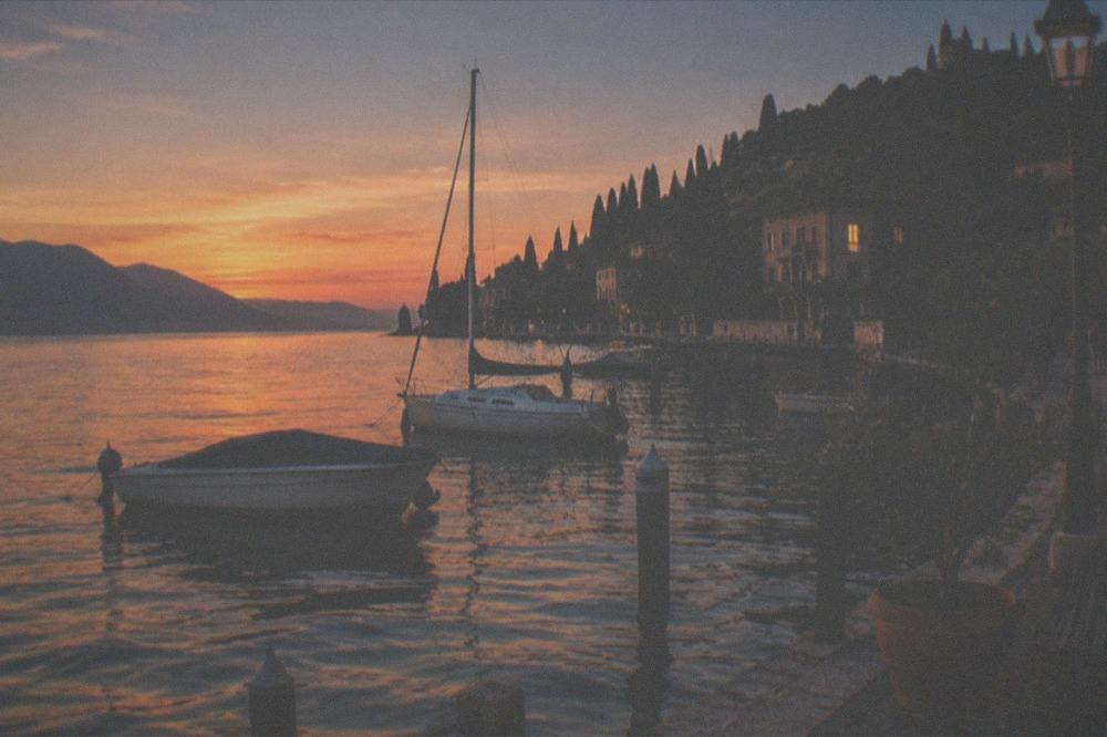 Moored boats on Lake Garda at dusk, orange light on the water, distant hills, and a tree-lined shore with buildings.