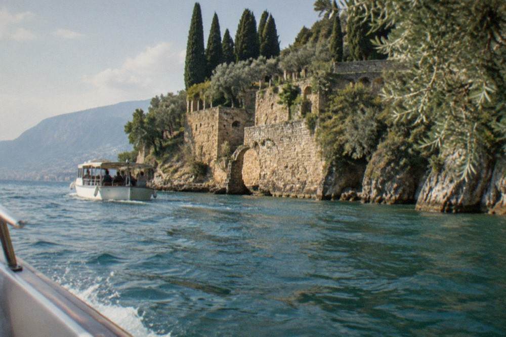 A small passenger boat passing beneath stone villa terraces above an olive grove on a lake headland