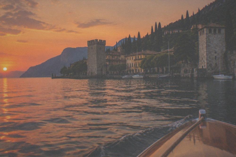 View from a wooden boat bow across Lake Garda toward a castle with towers and cypress trees on the shore at sunset.
