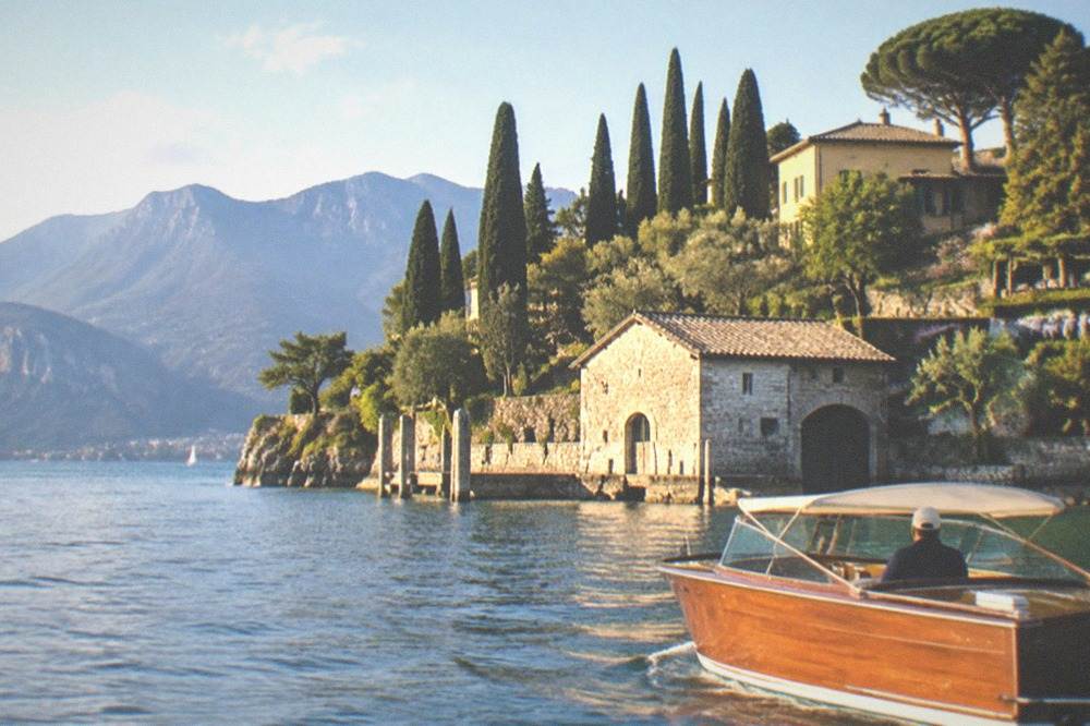 A wooden skippered boat gliding past a lakeside villa with cypress trees