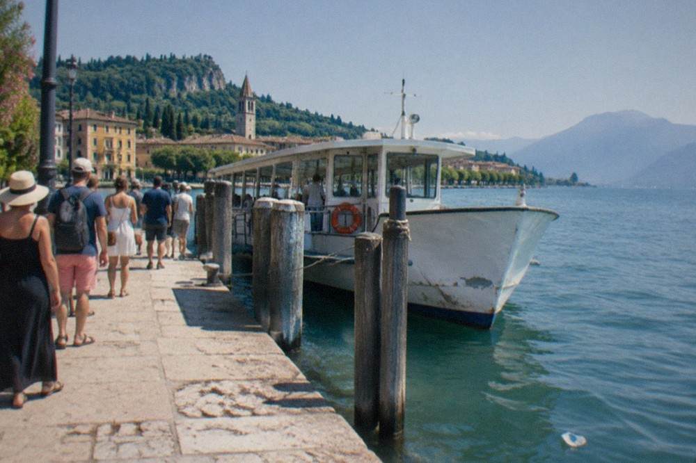 A busy stone landing stage on Lake Garda with a passenger boat tied up and a queue of visitors waiting in the sun
