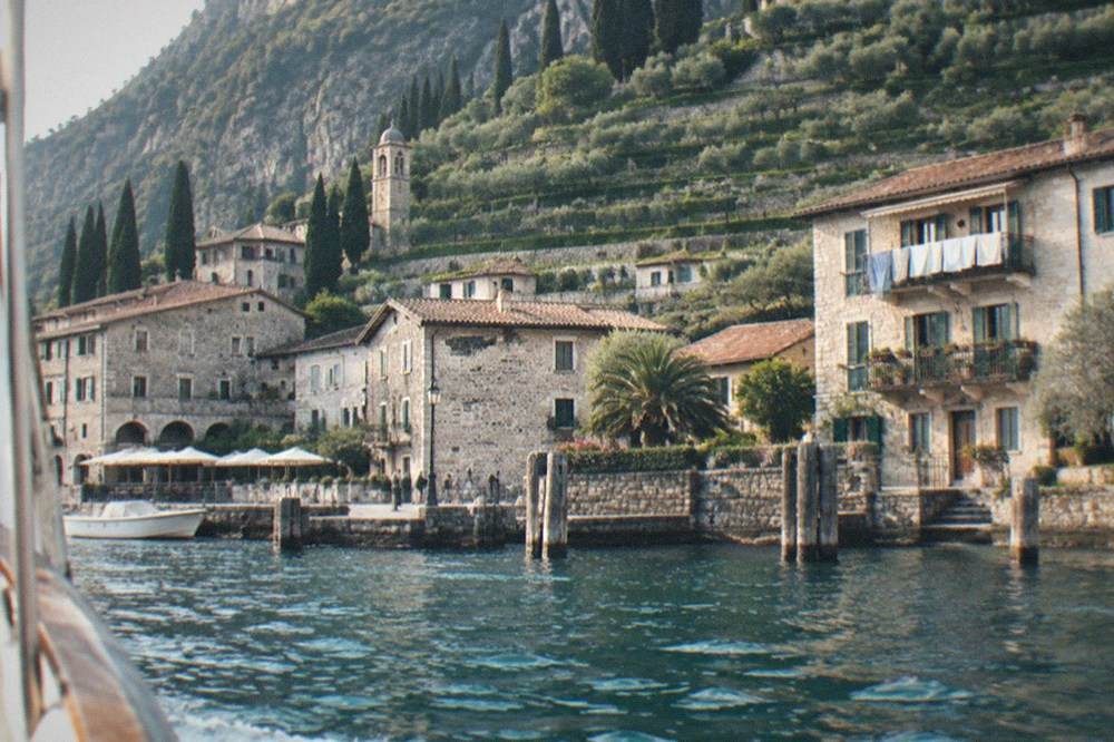 Stone Lake Garda lakeside houses, a church tower and a small harbour with a docked boat, terraced hills and cypresses behind, viewed from the water.