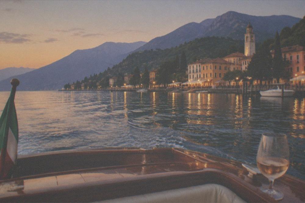 A wooden boat crossing calm evening water on Lake Garda with a lit town and hills on the far shore