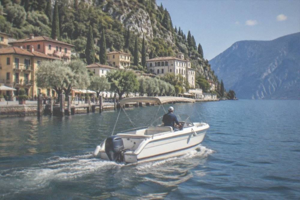 A small white motorboat with a person at the helm moves along a Lake Garda lakeside town of houses and trees beneath steep hills.