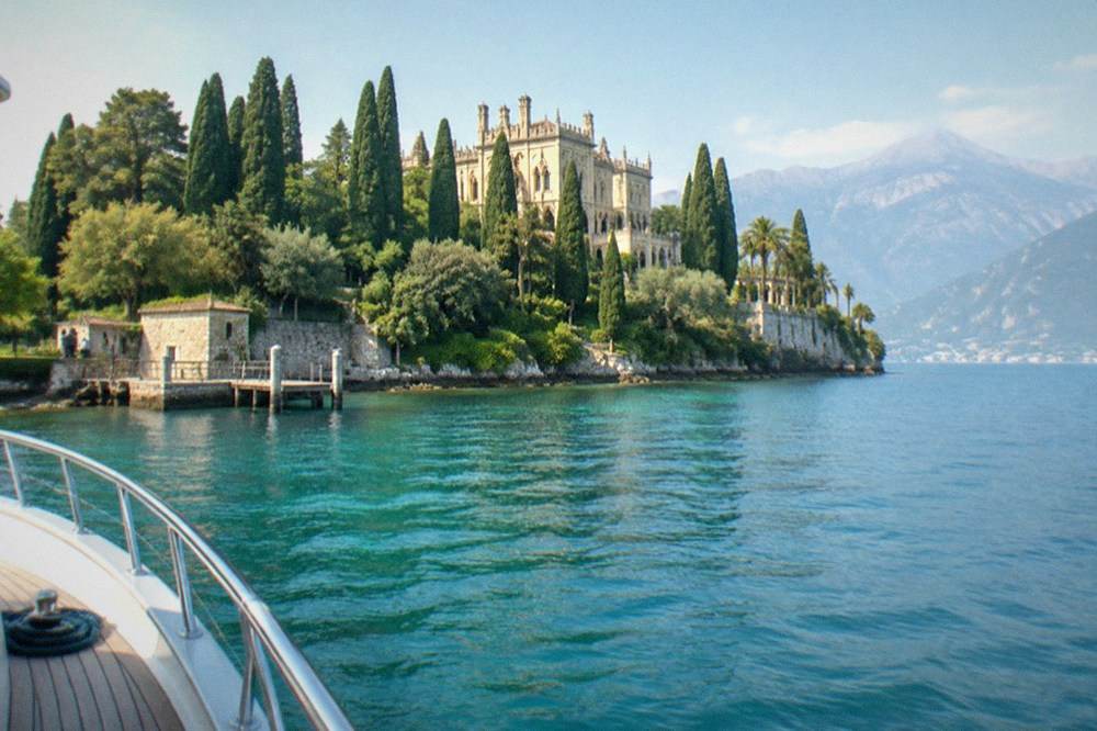 Ornate villa and tall cypress trees on a rocky lakeshore, seen from a boat bow over turquoise water with mountains beyond, on Lake Garda.