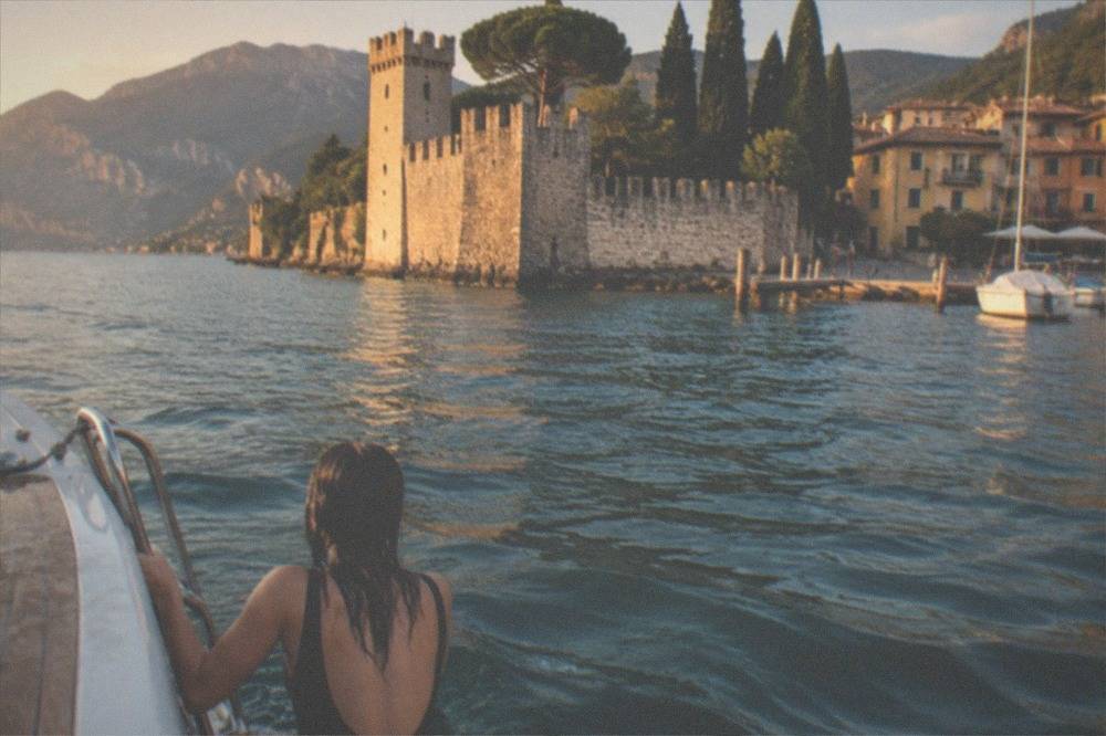 A person in a swimsuit sits on a boat facing a Lake Garda lakeside stone castle, cypress trees and mountains.