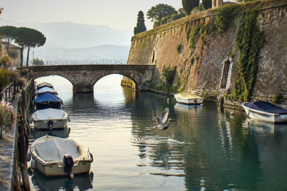 The walled old town of Peschiera del Garda seen across its moat with boats moored along the canal