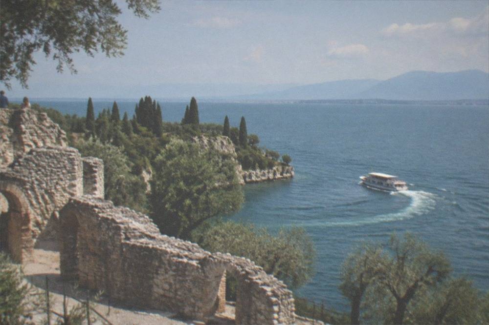 A small cruise boat rounding the Sirmione peninsula past the rocky headland and olive trees