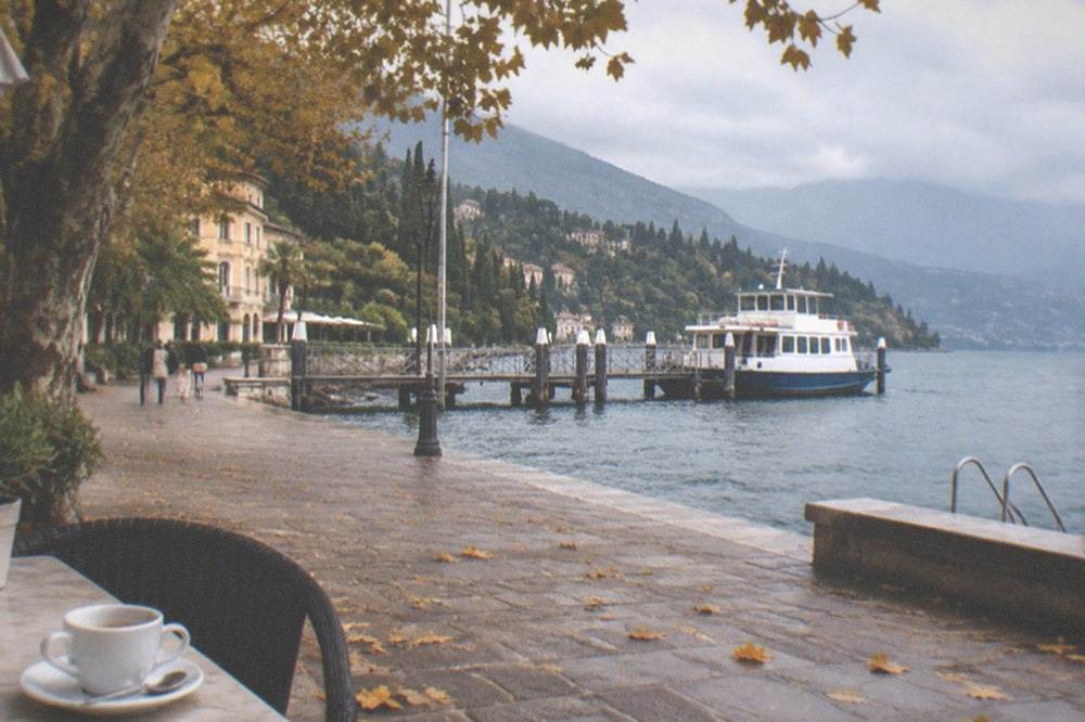 Lakeside stone promenade in autumn with a coffee cup in the foreground, a white-and-blue boat at a pier, walkers, and mountains beyond, on Lake Garda.