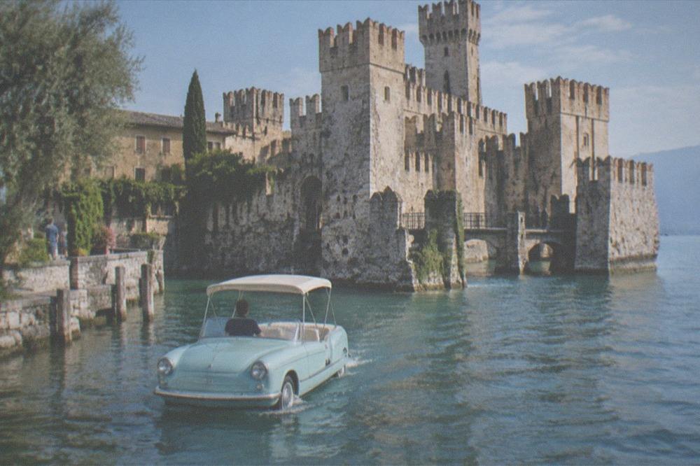 A pale blue amphibious car with a white canopy and driver moves on the water past a Lake Garda lakeside stone castle with battlements and towers.