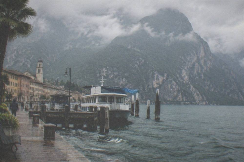 The northern basin of Lake Garda with a small town at the water's edge beneath steep mountain walls rising into cloud