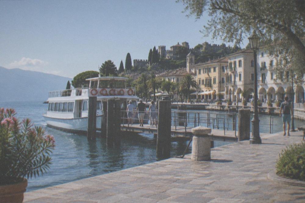 A guided tour boat moored at a Lake Garda landing stage with passengers boarding and a walled old town behind