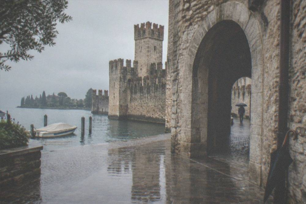 Stone Lake Garda lakeside castle with crenellated towers, an arched gateway, wet pavement, a moored boat and a person with an umbrella walking through.