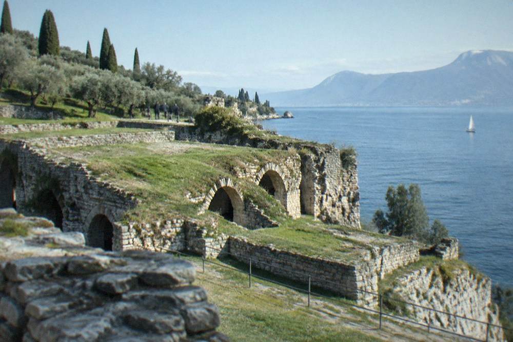 The stone walls of a Roman villa ruin above a lake with hills in the distance