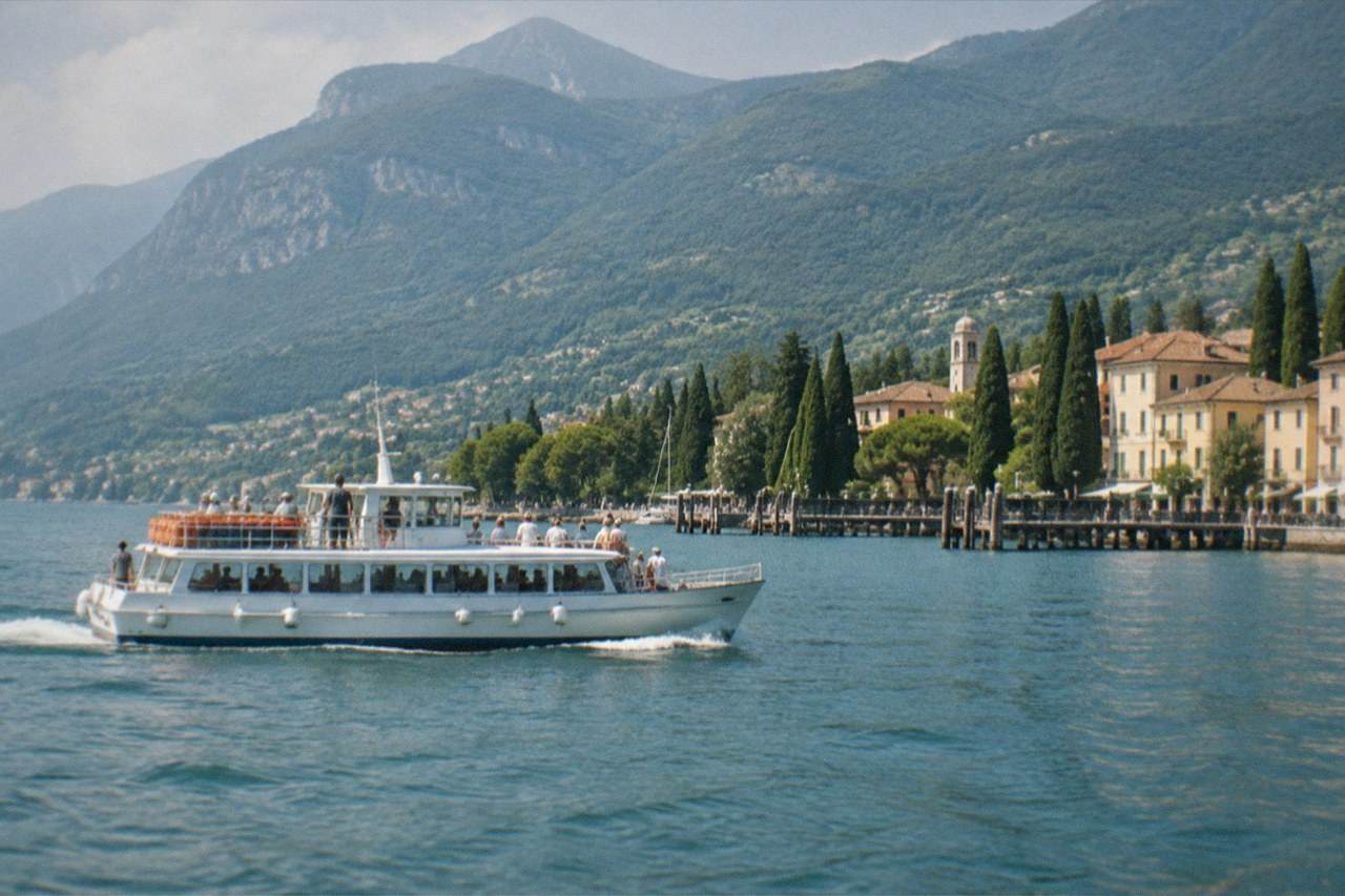 A white passenger boat with people on deck moves across Lake Garda, mountains and lakeside buildings behind it.