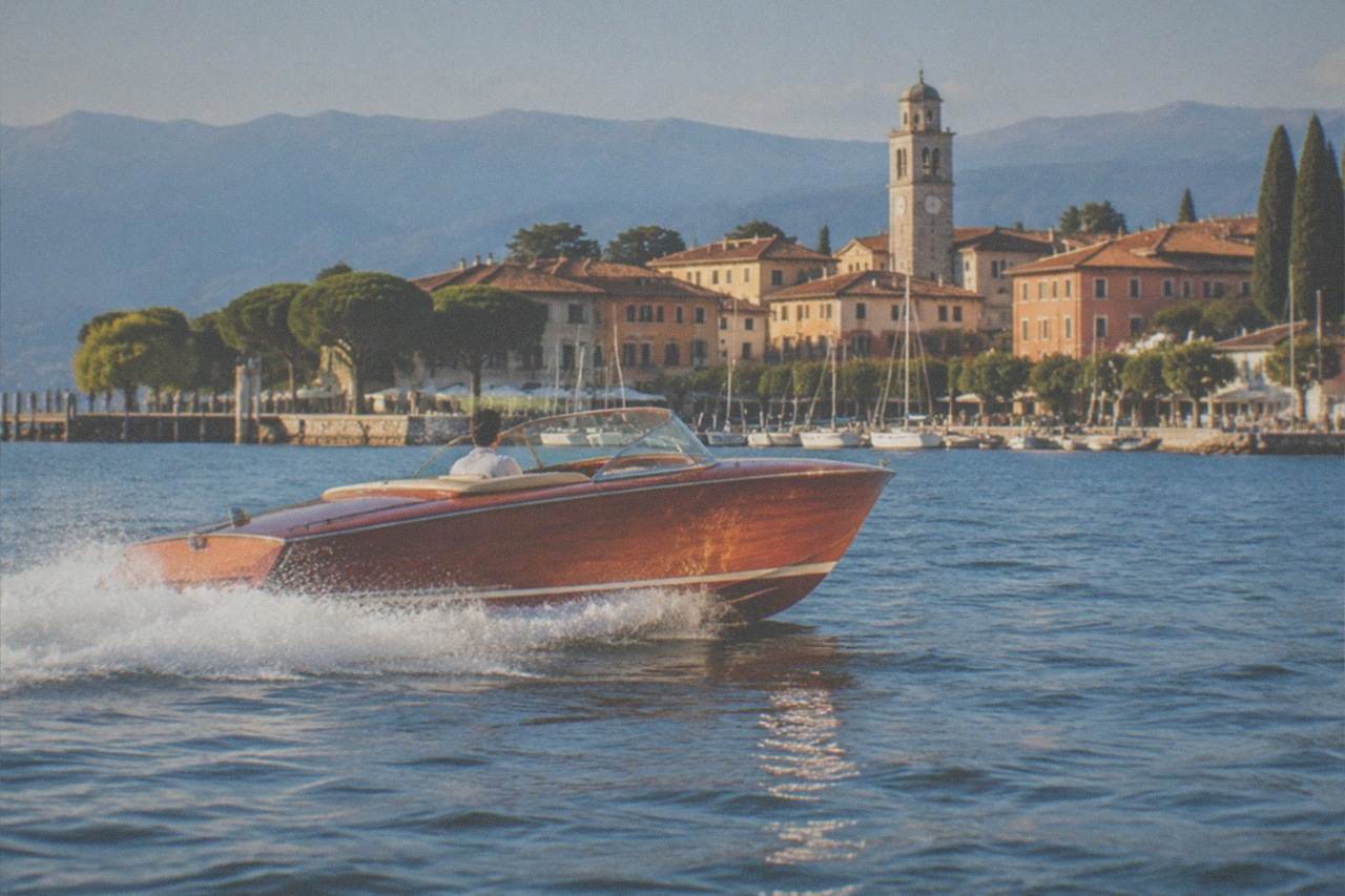 A varnished wooden runabout throws a white wake on Lake Garda, a driver at the wheel, with a clock-tower town, pines and mountains behind.